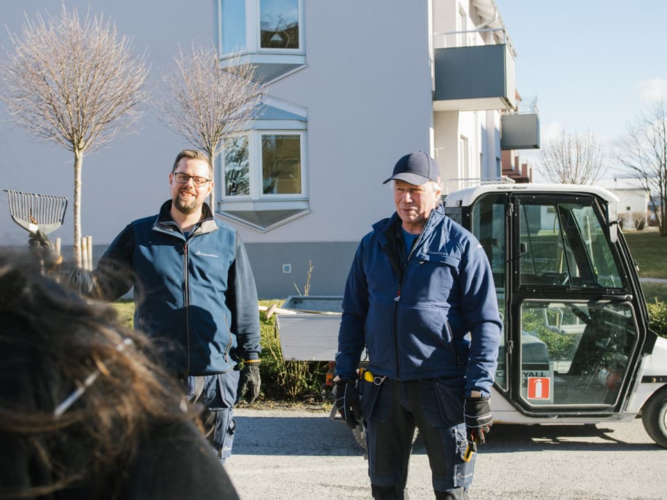 Två män med blåa jackor står utomhus, de tittar in i kameran och ler. I bakgrunden står en golfbil och ett vitt hus.
