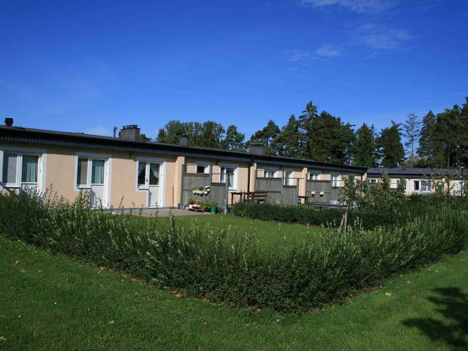 Avlångt orange enplanshus med uteplatser som delas upp med träplank. Framför huset är gräsmatta och låg häck. Himlen är blå.