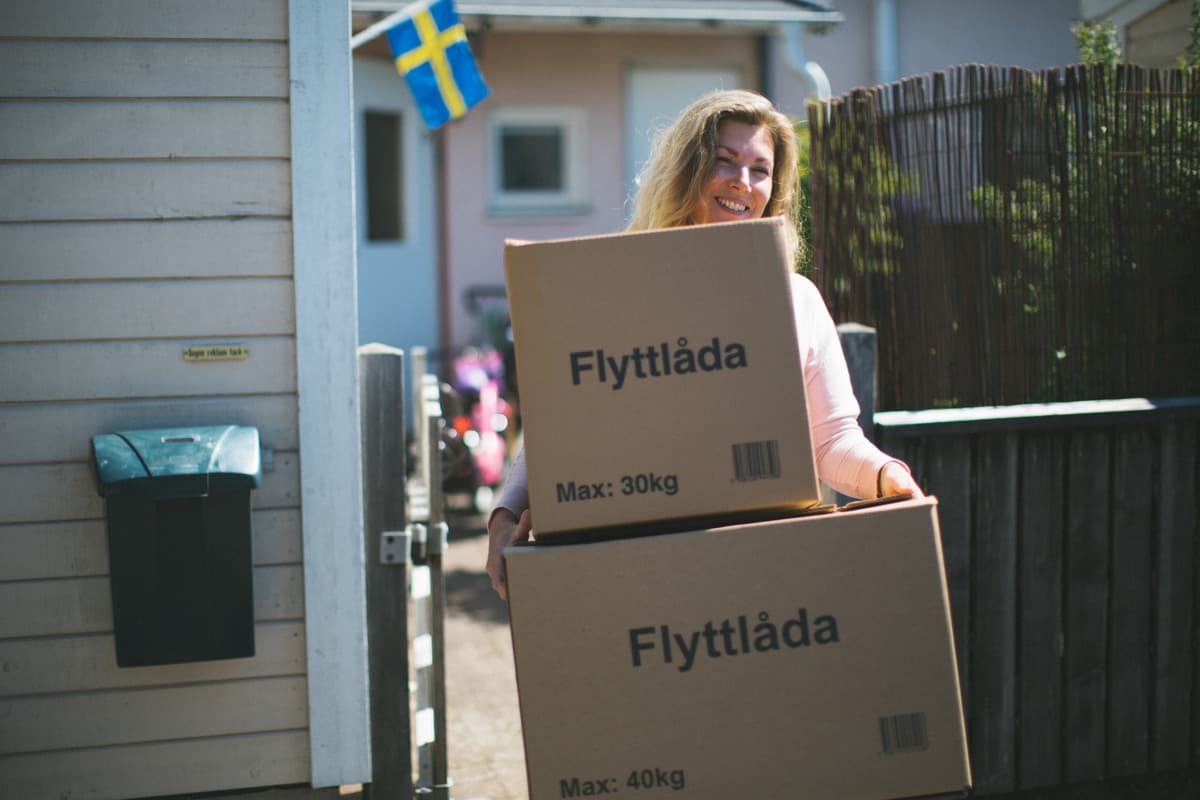 En kvinna framför ett radhus bär på två flyttkartonger.