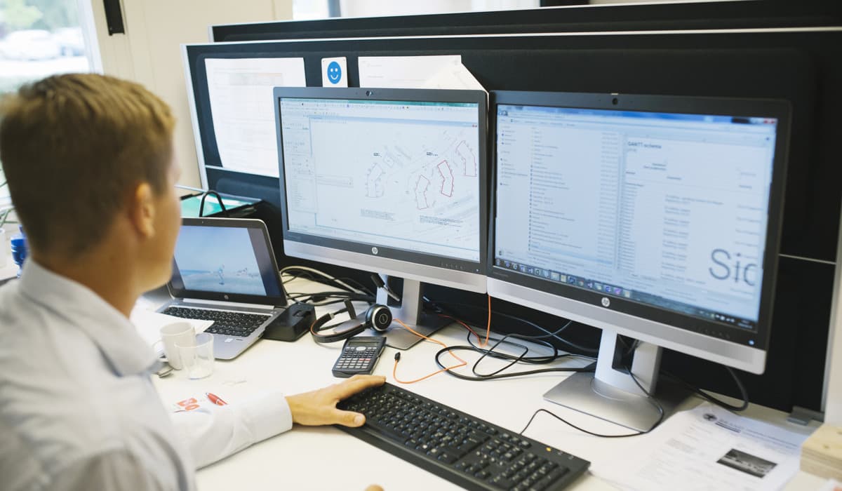 En man med ljusblå skjorta sitter vid ett skrivbord på ett kontor och kollar på en dator.