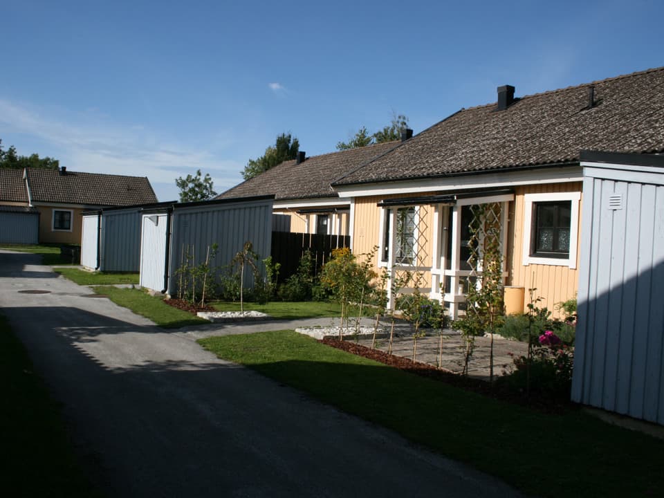 Gula radhus med vita knutar och separata ljusblå förråd. Framför husen är det gräsmatta och asfalterade stigar mellan smal väg och entré. Himlen är blå med ett fåtal moln.