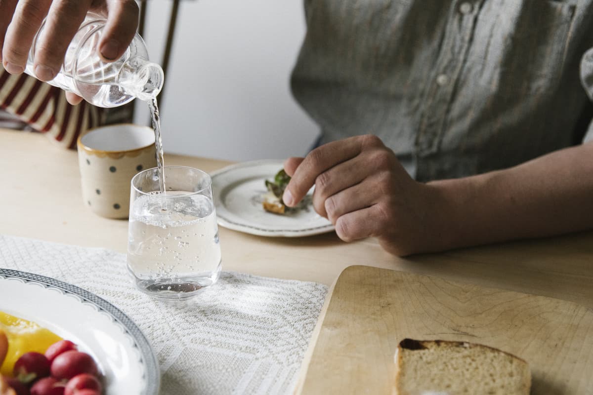 En person sitter vid ett bord och häller vatten från en karaff till ett glas. På bordet finns en vit och beige mönstrad duk, två tallrikar och en skärbräda i trä. Det är bara personens händer och armar som syns.