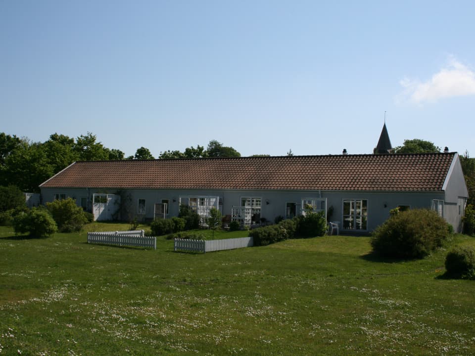 Avlångt grått hus med tegeltak. Framför huset är en gräsmatta och flera buskar. I bakgrunden syns ett kyrktorn. Himlen är ljusblå med ett moln.