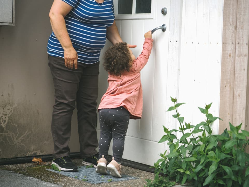 En kvinna står med en flicka vid en ytterdörr. Flickan sträcker sig för att öppna dörren.