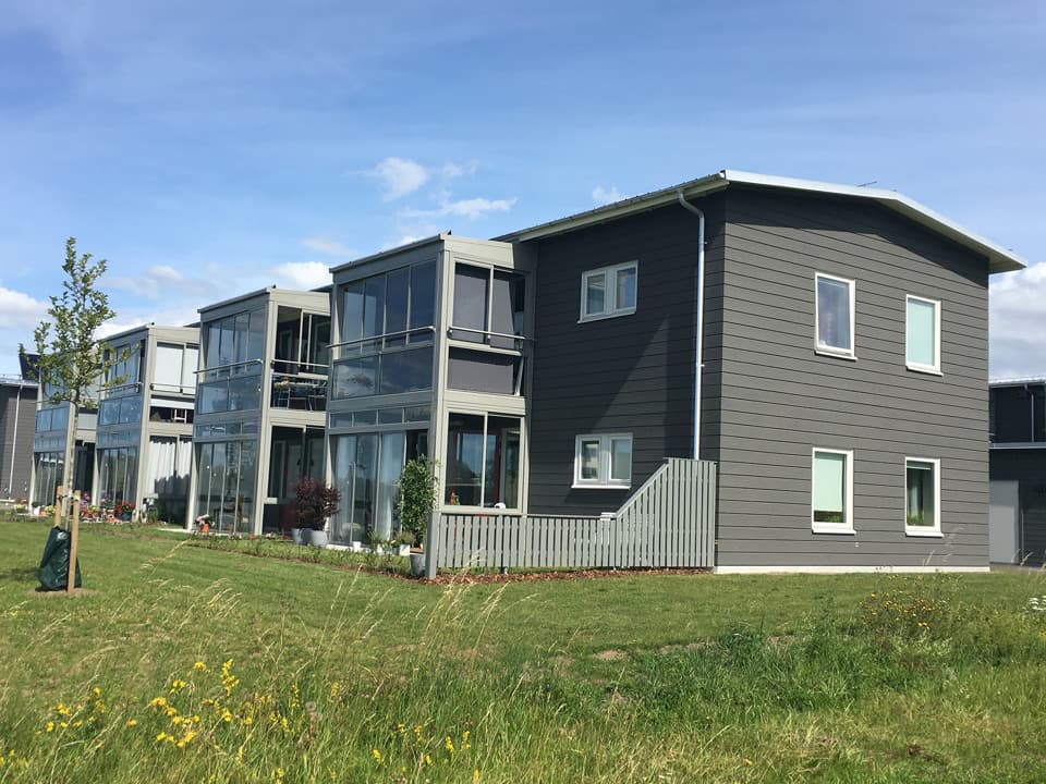 Mörkgrått tvåvåningshus med ljusgråa fönsterkarmar och ljusgråa inglasade balkonger. Framför huset är grön gräsmatta och himlen är blå med några få moln.