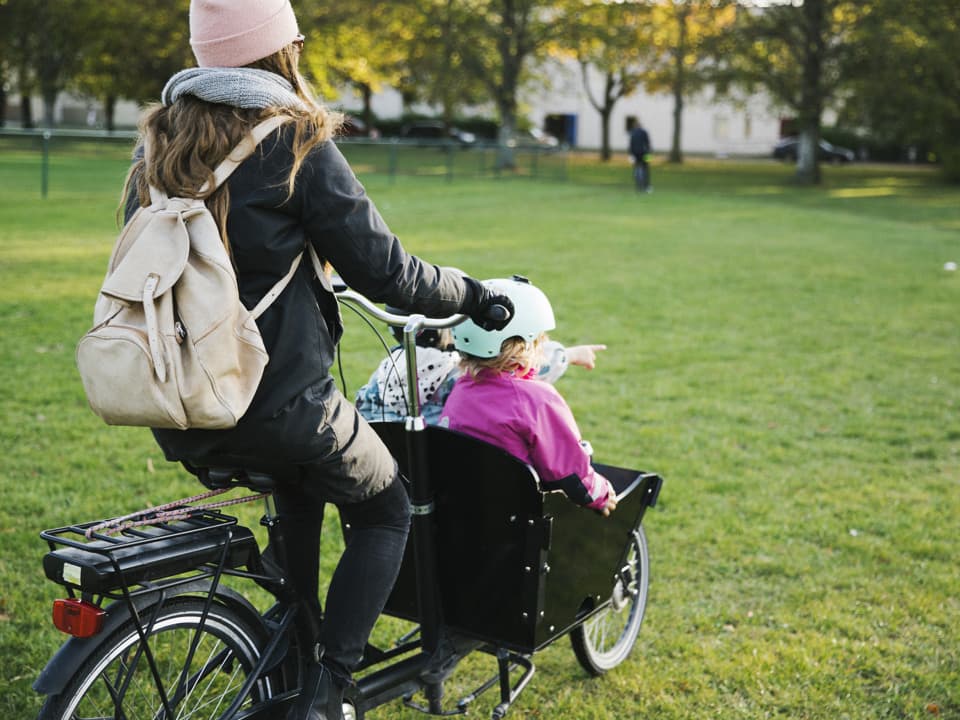 En kvinna kör två barn i en lådcykel på en gräsmatta. I bakgrunden är det hus och träd.