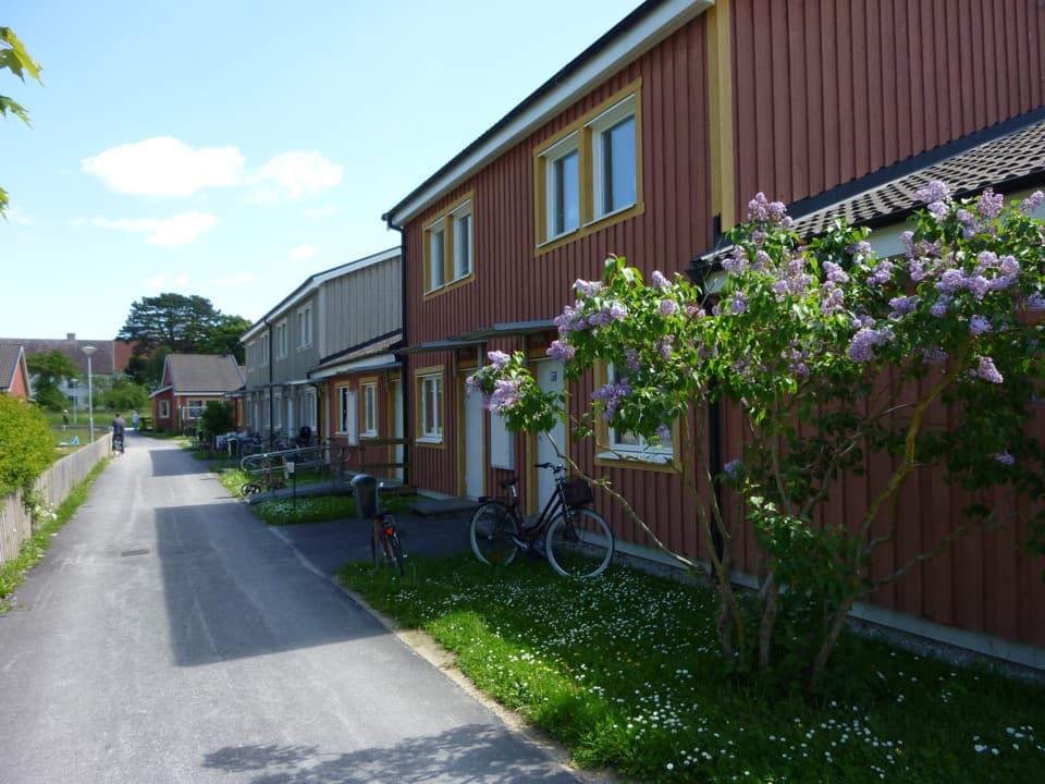 Rött trähus med gula knutar. Framför huset är en gräsplätt och därefter gång- och cykelväg. På gräset står en syrenbuske. 