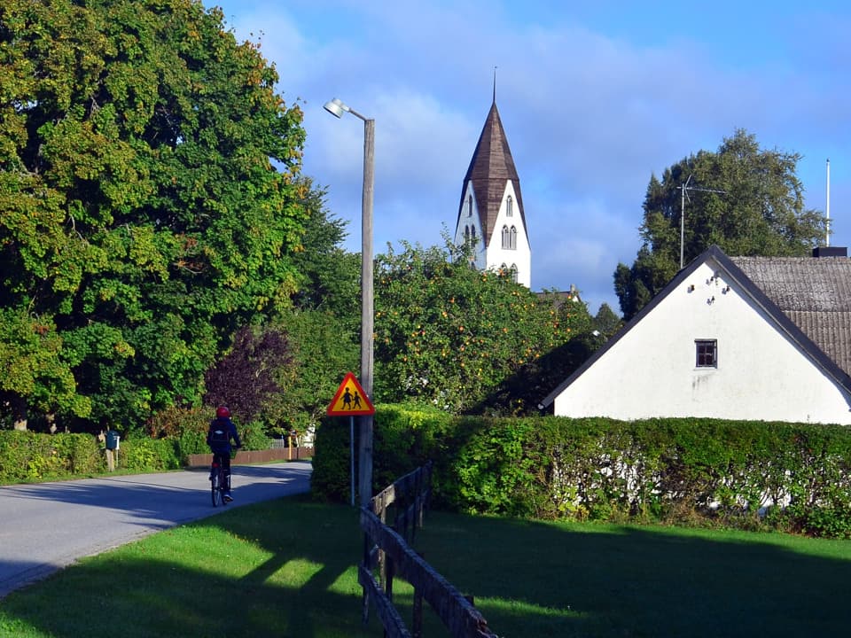 En person cyklar på en bilväg, längs med vägen är det grönt gräs, buskar och trä. Bakom en häck är ett vitt hus. Längs bort syns ett kyrktorn.