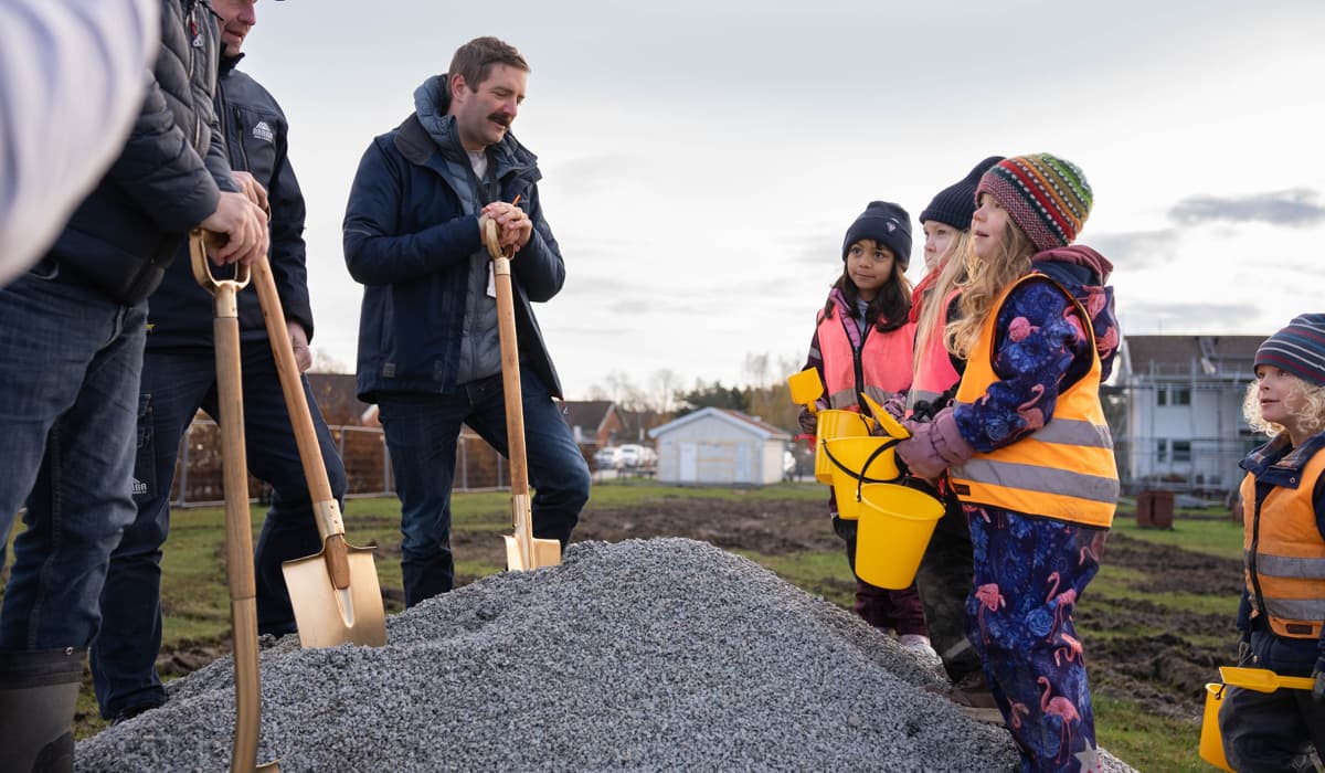Tre män i 40-50-årsåldern står på en grushög med varsin förgylld spade i händerna. Mittemot dem står fyra femåringar med gula plasthinkar och spade.