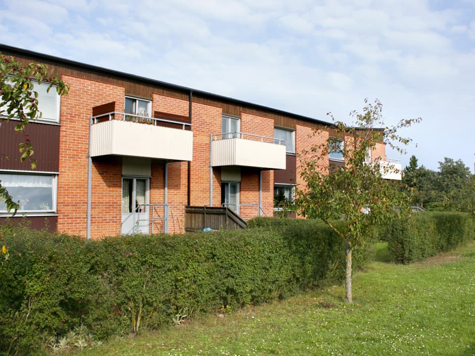 Rött flerfamiljs tegelhus med vita balkonger. Framför huset är det en häck, träd och gräsmatta.