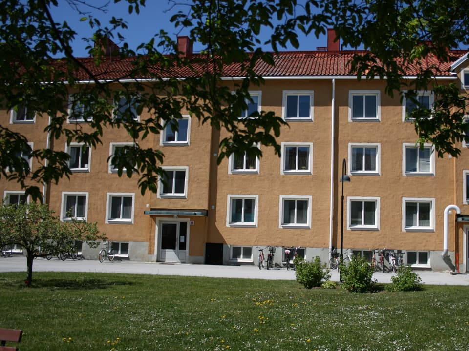Ett gult trevåningshus med vit fönsterkarmar och rött tegeltak. Framför huset går en smal, asfalterad cykel- och gångbana och framför den är en grön gräsmatta. I förgrunden syns löv på en trädgren.
