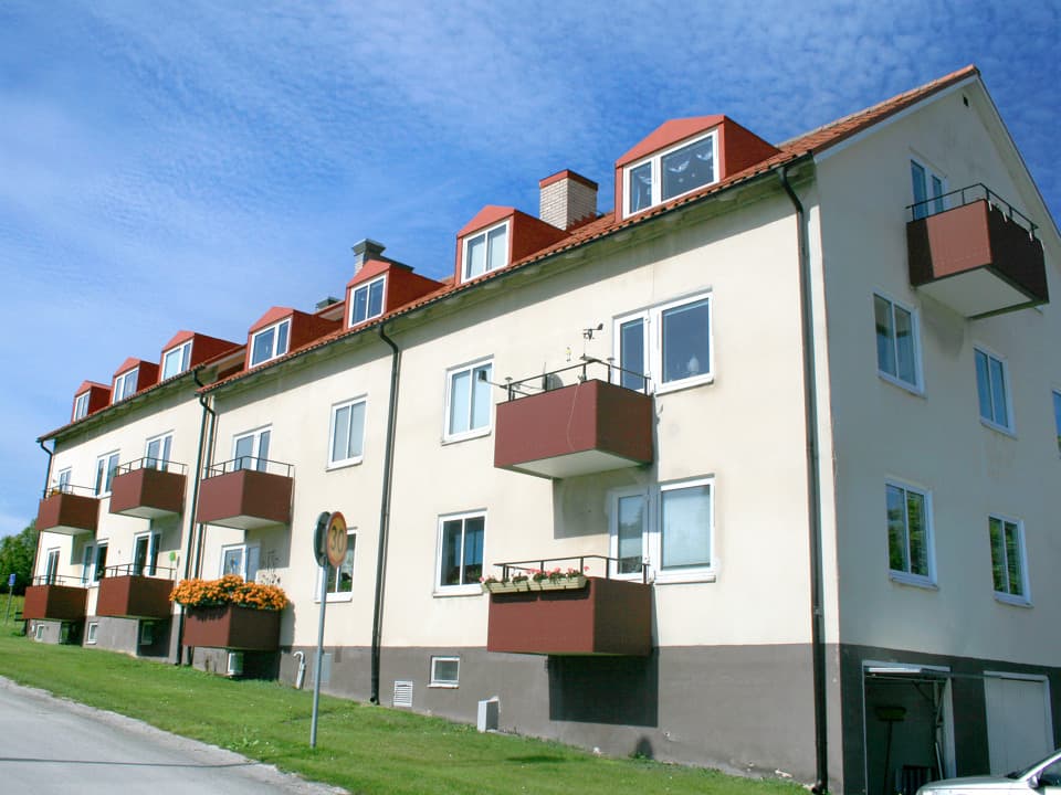 Vitt bostadshus med röda balkonger och rött tak. Framför huset är en gräsplätt. Framför gräset går en bilväg.