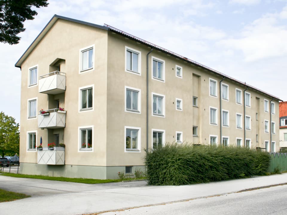 Treplans bostadshus med beige fasad, vita balkonger och vitt runt fönster. Framför huset går en häck och en trottoar. Det är flera moln på himlen.