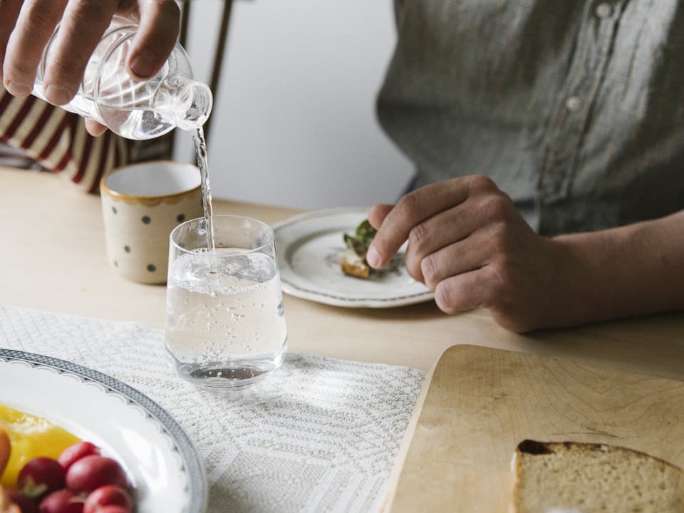 En person sitter vid ett bord och häller vatten från en karaff till ett glas. På bordet finns en vit och beige mönstrad duk, två tallrikar och en skärbräda i trä. Det är bara personens händer och armar som syns.