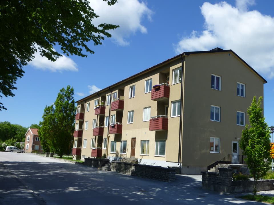 Gult bostadshus med röda balkonger. Framför huset går en asfalterad bilväg. Runt huset är några träd. Himlen är blå med vita moln.