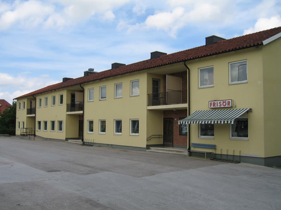 Ett långt gult bostadshus med två plan. På den del av huset som är närmast kameran finns en skylt där det står "Frisör".  Framför huset är det asfalterat, himlen är blå med flera moln.