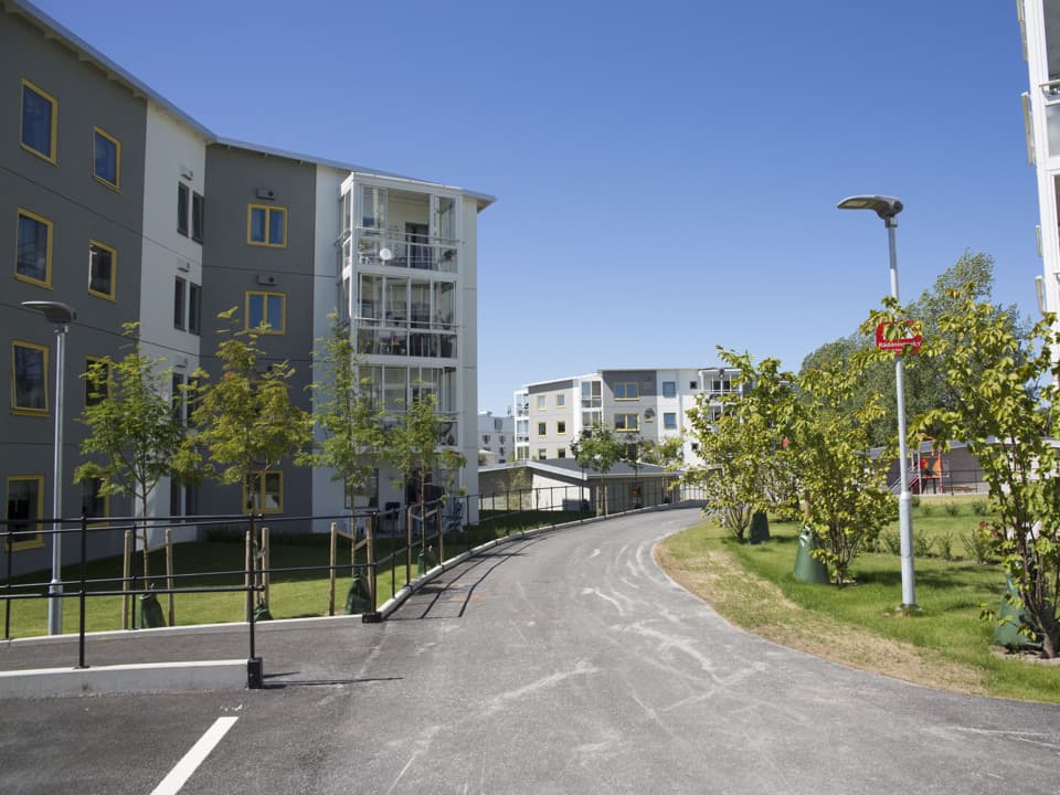 En asfalterad väg går genom ett bostadsområde. Mellan husen och vägen är det grön gräsmatta och små planterade träd. Husen har grå och vit fasad. Himlen är blå.