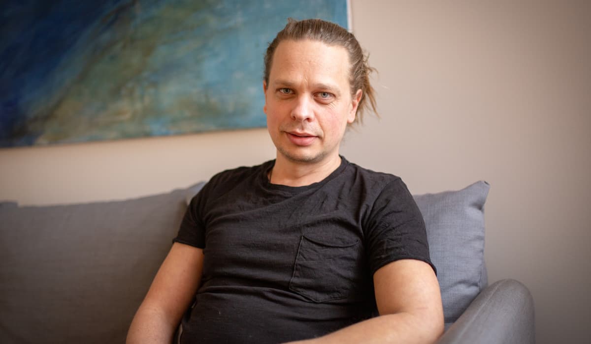 En man sitter i en grå soffa. Han tittar in i kameran och ler. På väggen hänger en tavla med blåa nyanser.