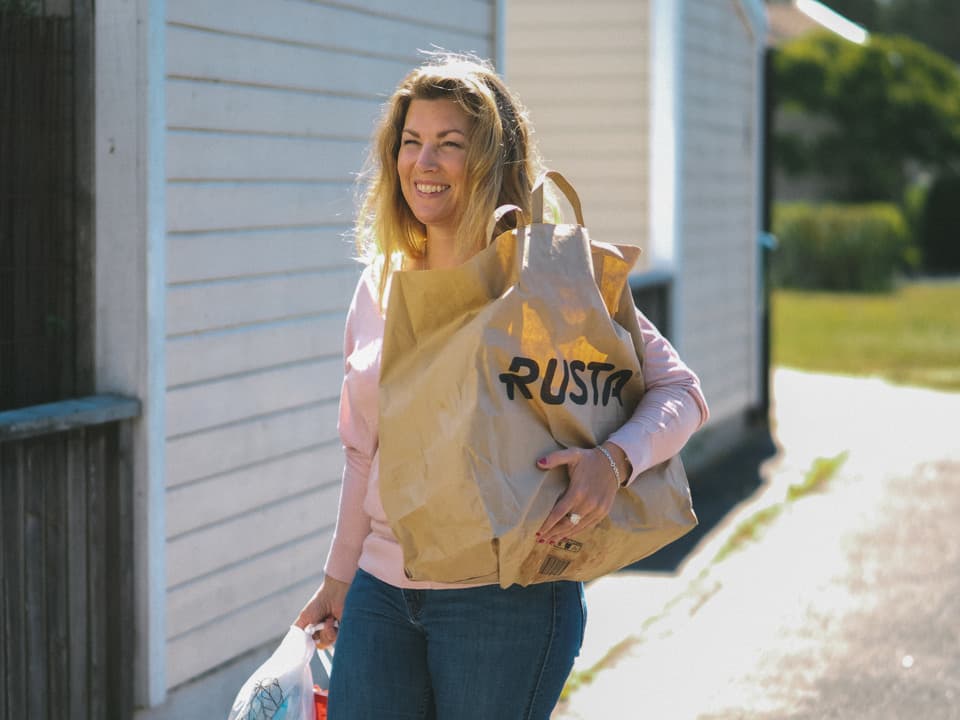 En kvinna med rosa tröja och jeans går i ett bostadsområde utomhus, hon bär på påsar.