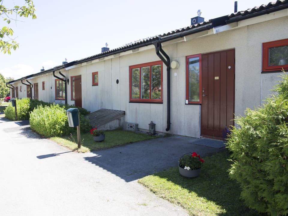 Avlångt flerfamiljshus med vit fasad och röda fönsterkarmar och ytterdörr. Framför huset är gräsplätt och en asfalterad väg. Himlen är ljusblå.
