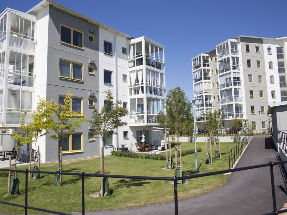 Två bostadshus bredvid varandra. Båda husen har grå och vit fasad, vita och inglasade balkonger och gula fönsterkarmar.