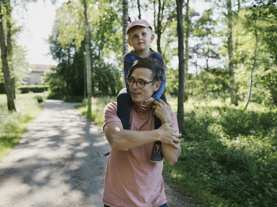 En pappa har sitt barn på sina axlar, de går på en väg med träd och buskar på vardera sida.