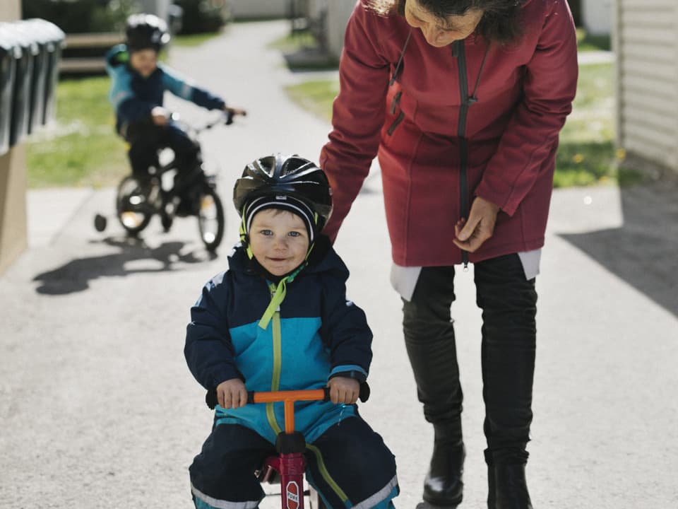 En pojke med hjälm på huvudet cyklar. En kvinna böjer sig ned mot pojken. I bakgrunden kommer en till pojke cyklandes. De är i ett bostadsområde.