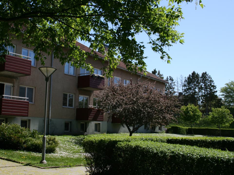 Flerfamiljshus med brunröd fasad och balkonger. Framför huset är det gräsmatta, buskar och träd. Himlen är ljusblå.