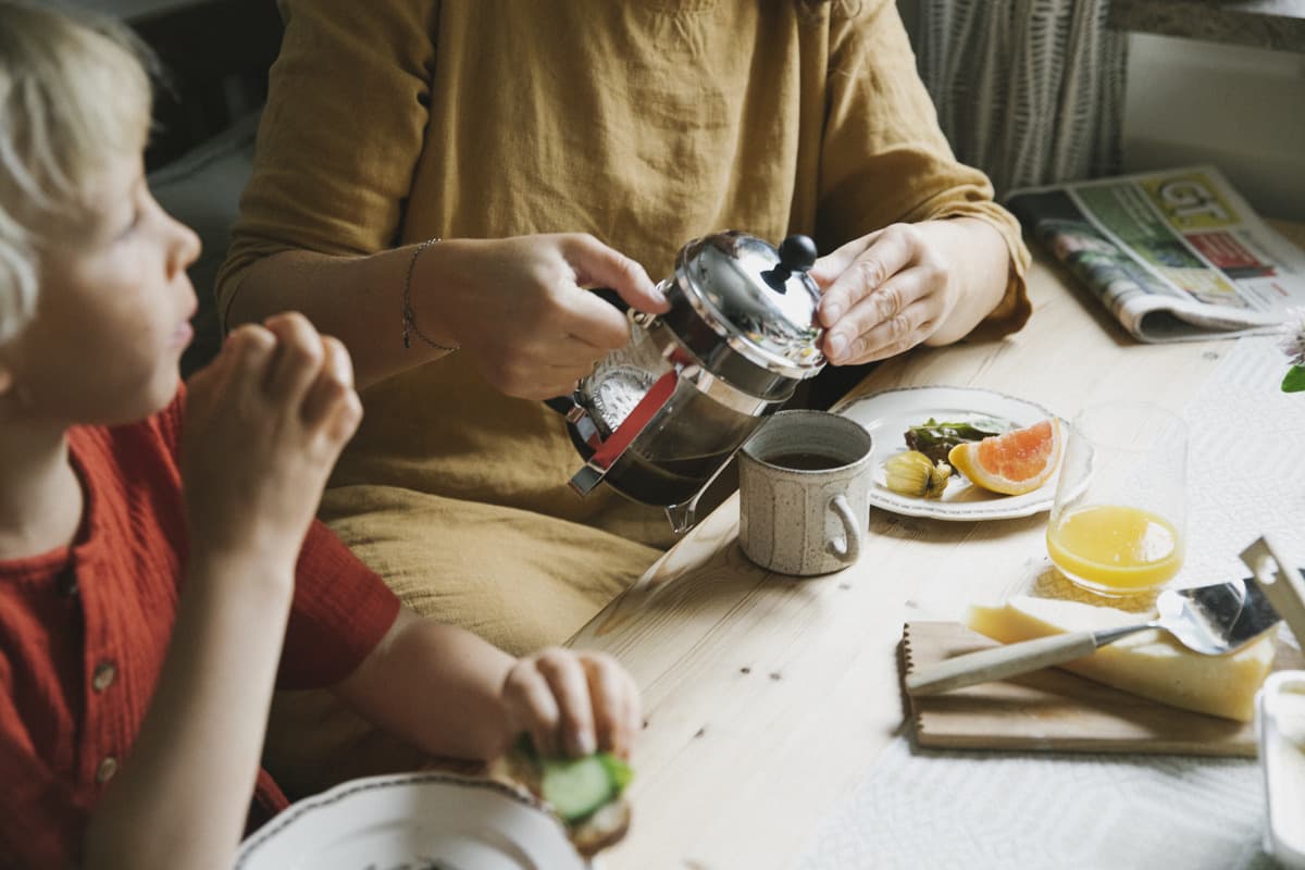En kvinna och en flicka sitter vid ett träbord, på bordet är det uppdukat för frukost. Kvinnan häller upp kaffe i en kopp. Flickan äter gurka.