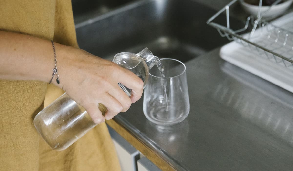 En kvinna med gul klänning står vid en diskbänk och häller vatten från en karaff till ett glas. 