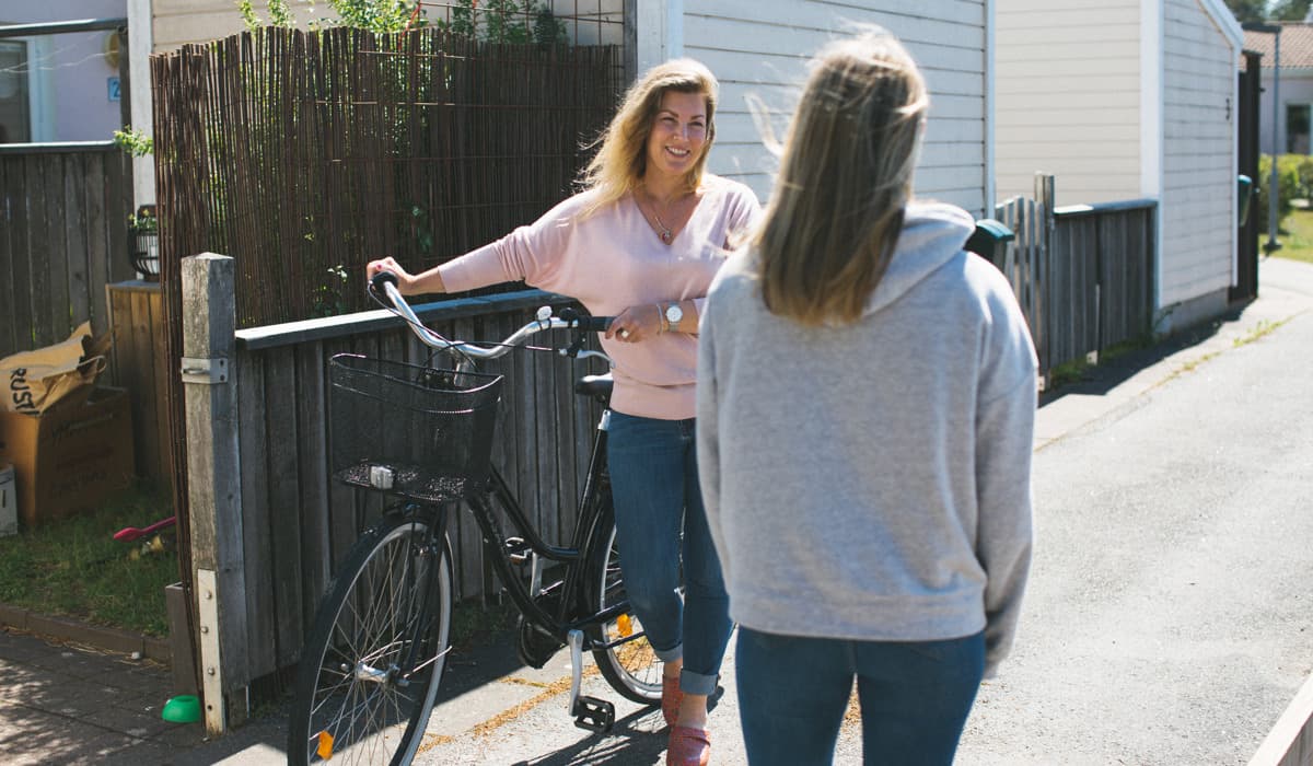 Två kvinnor pratar med varandra i ett bostadsområde. Den ena kvinna står med ryggen till och har en grå hoodie. Den andra kvinnan är vänd mot kameran, har en rosa tröja och håller i en cykel. 