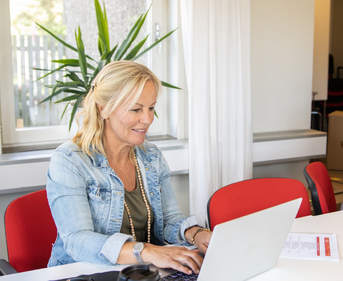 En kvinna med jeansjacka sitter inomhus vid ett bord och skriver på en laptop.