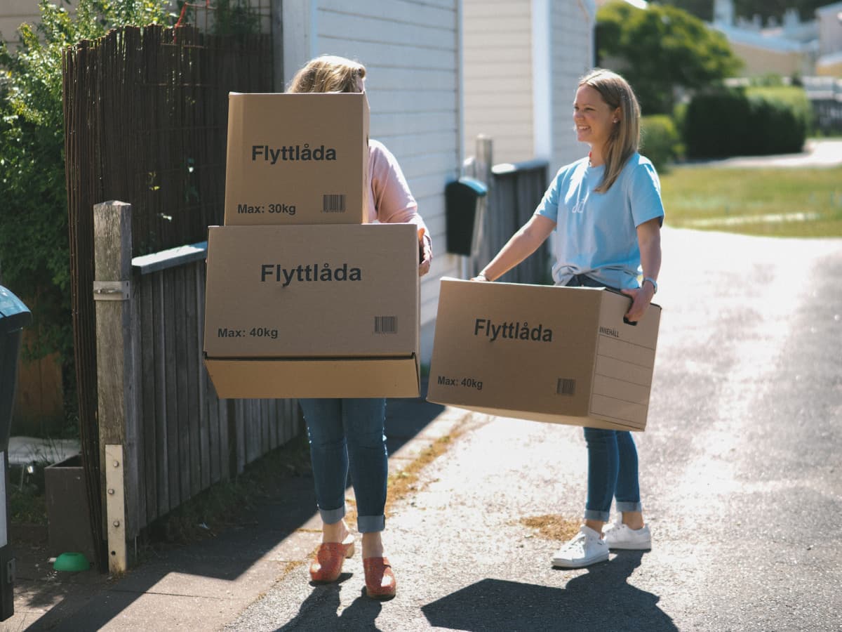 Två kvinnor bär på flyttkartonger. Den ena kvinnan har en rosa tröja och bär två kartonger, den andra kvinnan har en ljusblå t-shirt och bär en kartong. De är utomhus i ett bostadsområde.