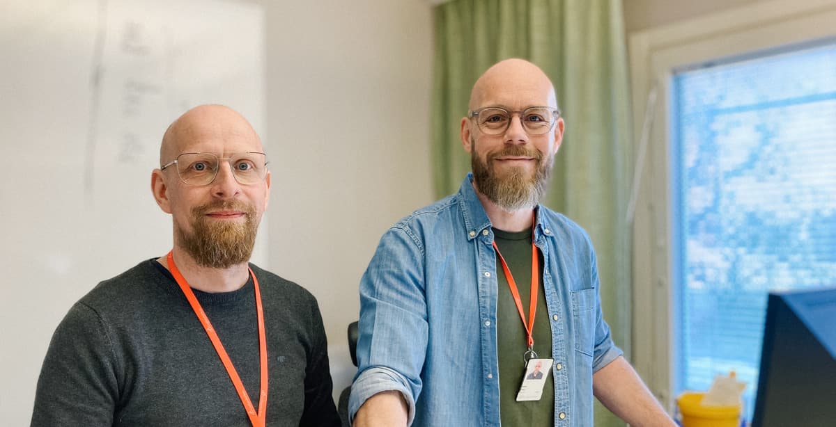 Två män med glasögon och skägg står framför en dator. De tittar in i kameran och ler.