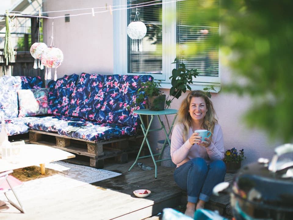 Kvinna ler glatt med en kaffekopp i händerna på sin uteplats.