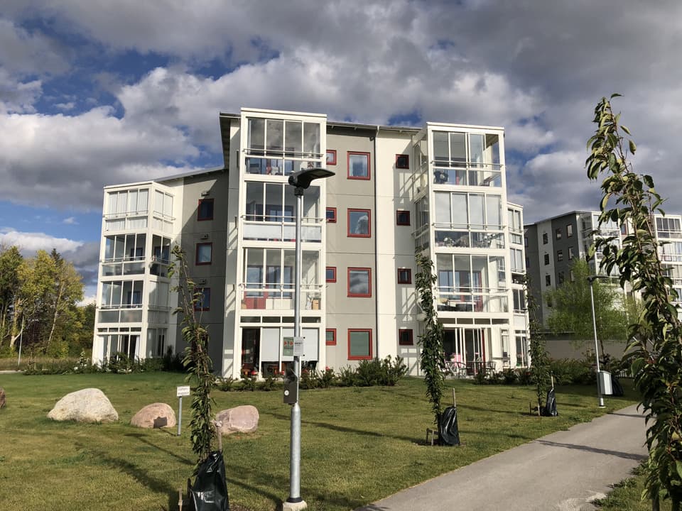 Ett fyra våningar högt bostadshus med grå och vit fasad, röda fönsterkarmar och vita, inglasade balkonger. Framför huset är det gräsmatta och himlen är blå med moln.