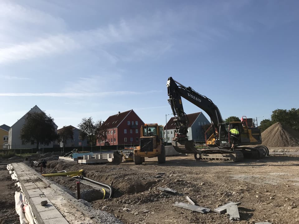 Två grävmaskiner gräver i marken för kvarteret Bläckfisken. I bakgrunden syns GotlandsHems andra kvarter, Castor. Det ligger en stor grushög bakom en av maskinerna. Himlen är blå. 