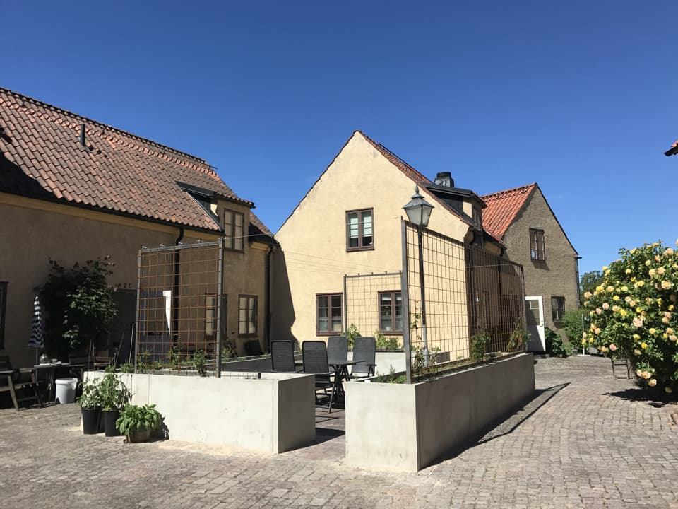 En pergola med betongrabatter, innanför den står ett svart bort med stolar. I bakgrunden är ett gult stenhus med bruna fönsterkarmar. Himlen är blå.