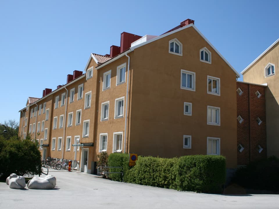Ett gult trevåningshus med vita fönsterkarmar.Framför huset går en smal, asfalterad cykel- och gångbana och på sidan en häck.