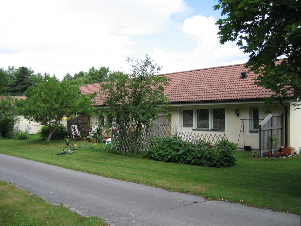 Enplanshus med ljusgul putsfasad och rött tegeltak. Längs huset finns två uteplatser separerade med ett plank. Framför dem är det gräsmatta och en smal, asfalterad gångväg.
