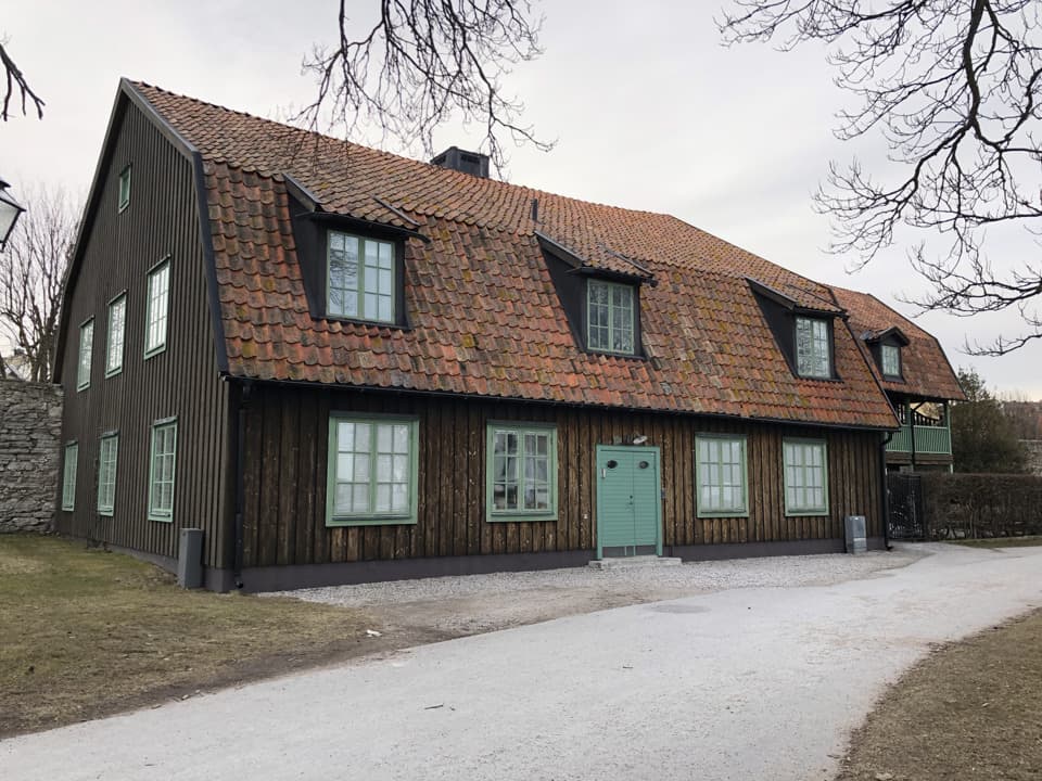 Ett trähus med brun fasad och gröna fönsterkarmar och dörr. Framför huset går en asfalterad väg. Det är moln på himlen.