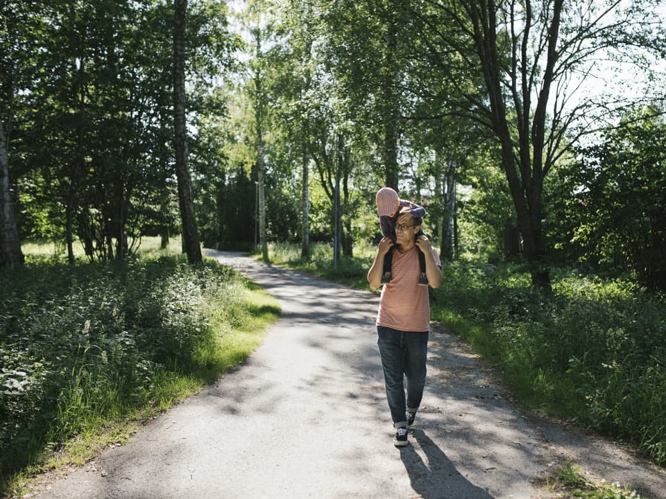 En pappa har sitt barn på sina axlar, de går på en stig genom en skogsdunge.