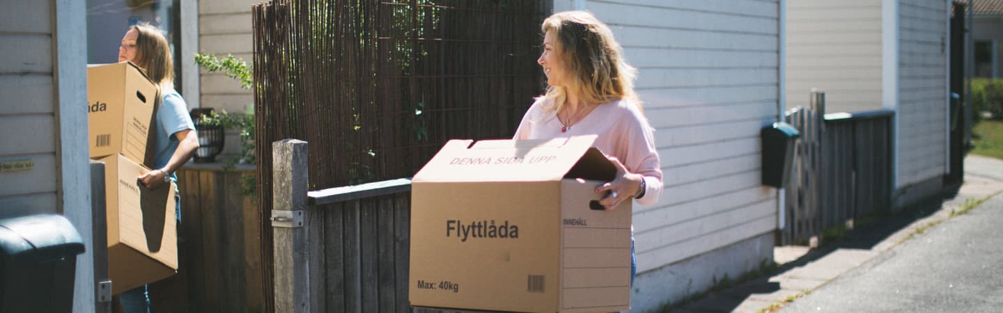 Två kvinnor bär på flyttkartonger. Den ena kvinnan bär två kartonger, den andra kvinnan bär en kartong. De är utomhus i ett bostadsområde.