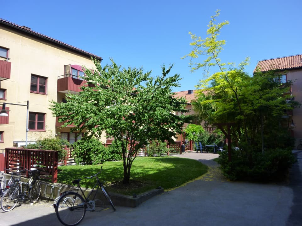 En innergård vid ett gult bostadshus. På innergården är en gräsmatta, en liten sittplats, träd och cykelställ. Himlen är blå.