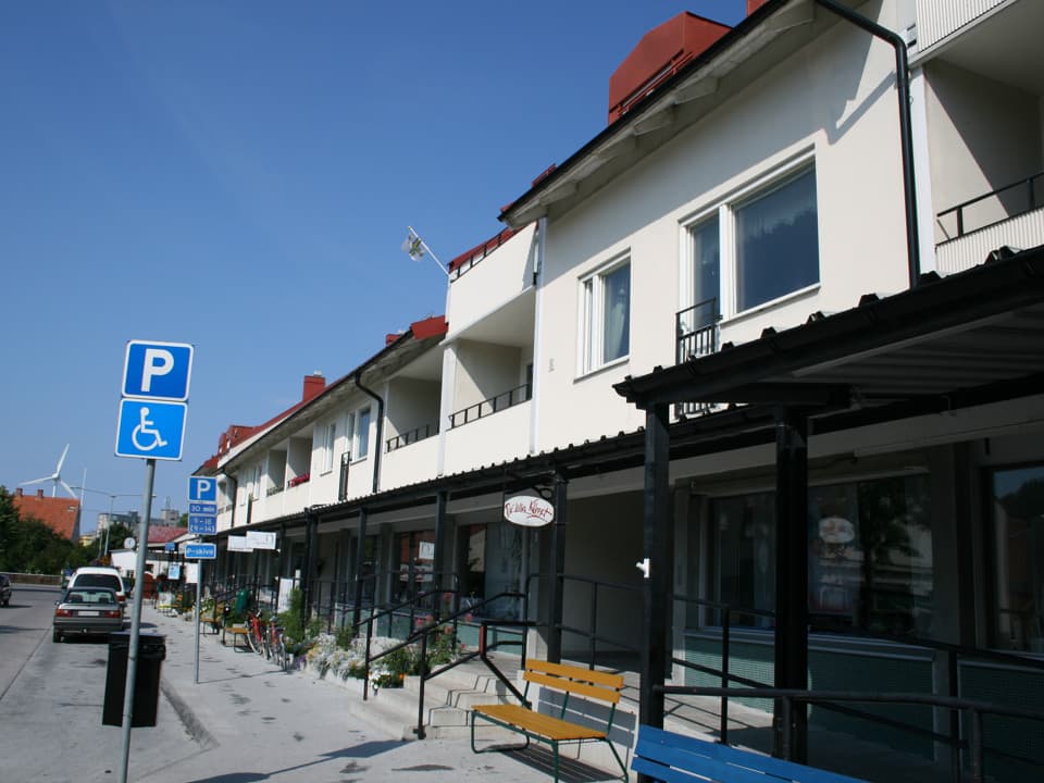 En vit byggnad med butiker och lokaler på bottenplan och bostäder på övre plan. Byggnaden ligger längs med trottoar och bilväg. Himlen är blå.