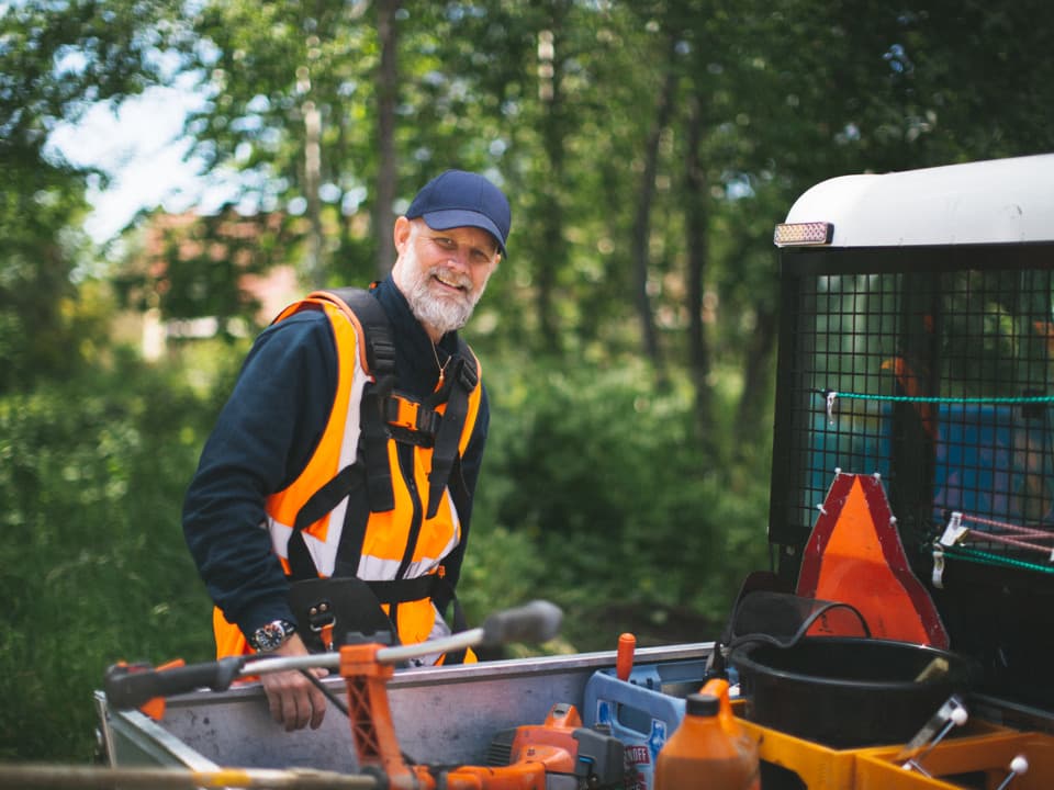 En man med svart keps, svart jacka och orange arbetsväst tittar in i kameran och ler. Han håller ena handen på ett bilflak. I bakgrunden är gröna träd och buskar.