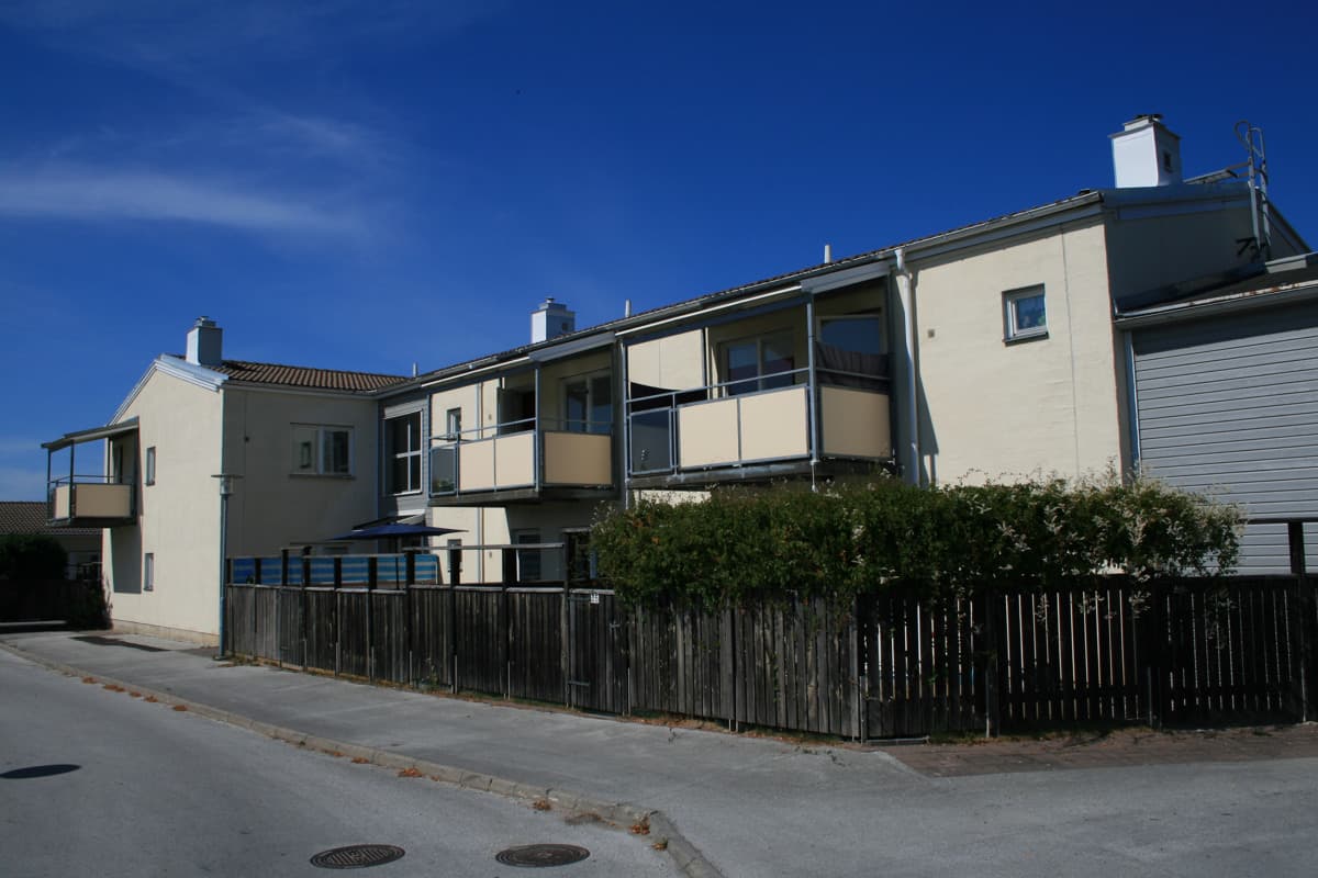 Ett vitt bostadshus med balkonger stor bakom ett träplank. Framför planket är en trottoar.