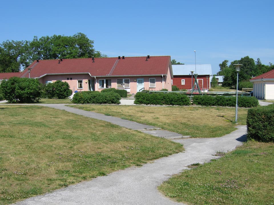 Ett rosa, ett rött och ett vitt hus står tillsammans i en klunga. Runtomkring är gröna buskar och gräsmatta. Genom gräset går smala asfalterade stigar. Himlen är blå.