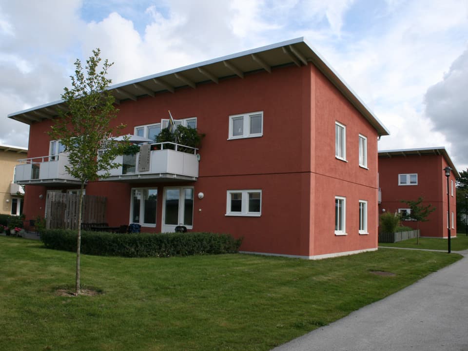Ett rött tvåplans bostadshus med vita fönsterkarmar. På övre plan är vita balkonger och på nedre plan en uteplats. Framför huset är det grön gräsmatta och ett litet planterat träd.