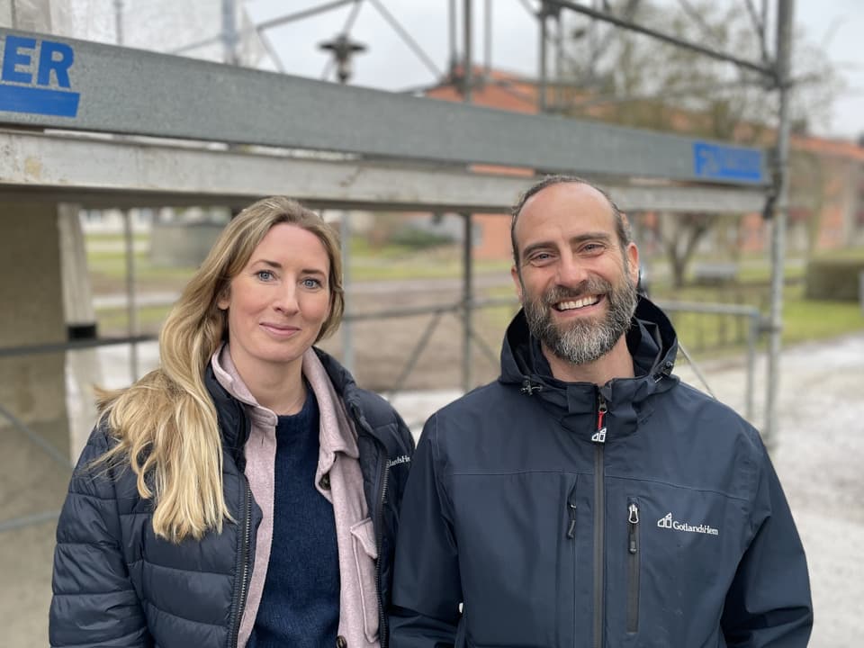 En man och en kvinna står framför en byggställning. De tittar in i kameran och ler.
